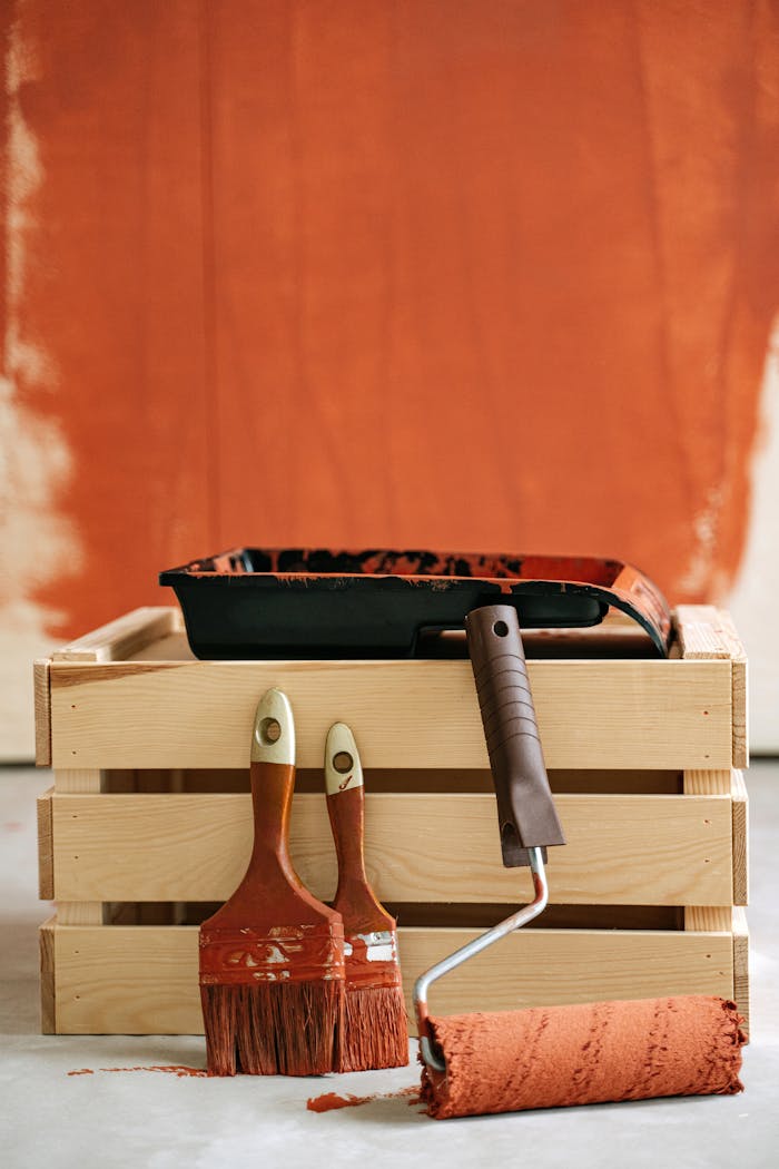 Wooden crate with paintbrushes and roller against a partially painted wall.